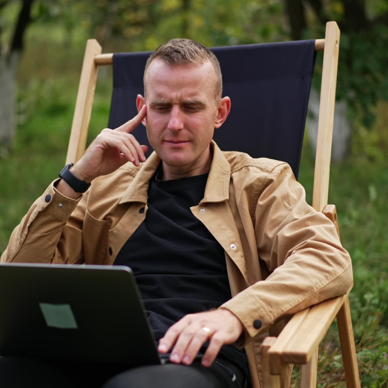 Pensive sitting in garden chair looks thoughtfully at his laptop screen. Freelancer working in nature has found some mistake and looks closer at computer