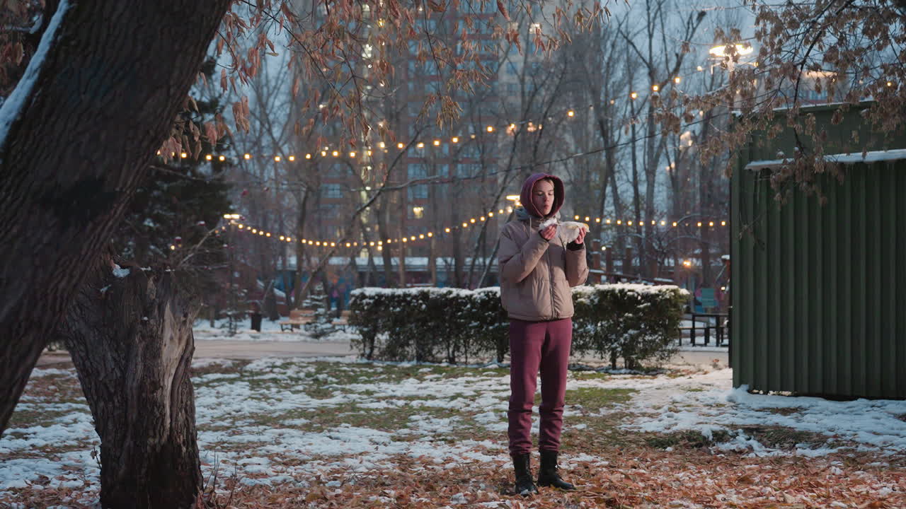 молодая женщина с салфеткой в руке и еда кукурузы, стоя в парке со снегом, праздничные огни на заднем плане, расслабленный момент на открытом воздухе, наслаждение едой