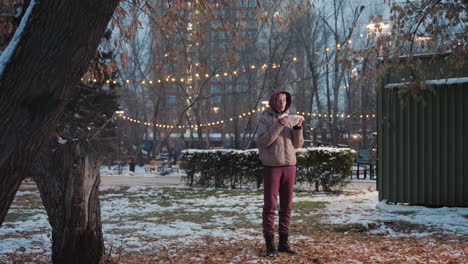 young woman holding napkin in hand and eating corn while standing in park with snow, holiday lights in background, relaxed outdoor moment, food enjoyment