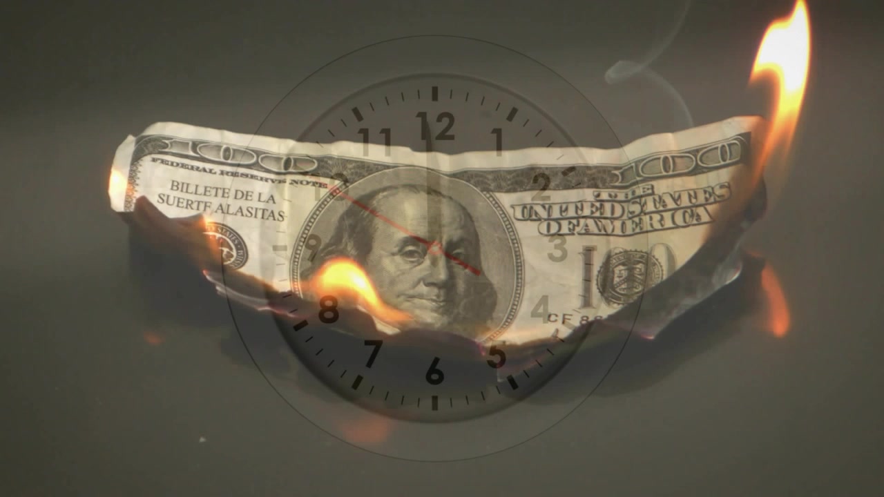 Piccola scintilla che accende l'estremità destra, banconota da 100 dollari che brucia e si arriccia, sovrapposizione di un orologio che mostra il conto alla rovescia