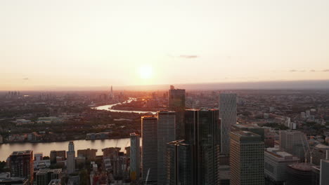 Aerial-panoramic-footage-of-skyscrapers-in-modern-business-borough-Canary-Wharf.-Thames-river-flowing-through-city.-View-against-setting-sun.-London,-UK