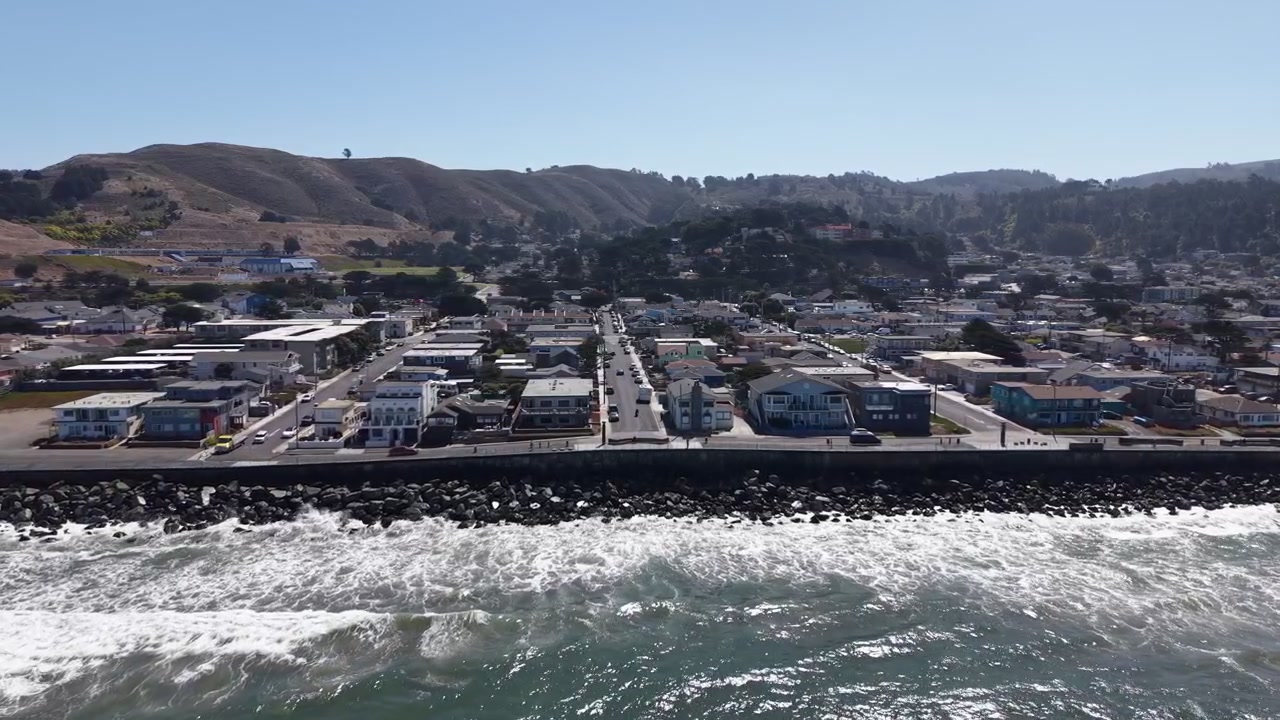 Una ripresa con drone rivela il quartiere fronte oceano di Pacifica, California, nella nitida luce del mattino. Le onde si infrangono contro la costa rocciosa, con colline illuminate dal sole e cieli azzurri e limpidi sullo sfondo.
