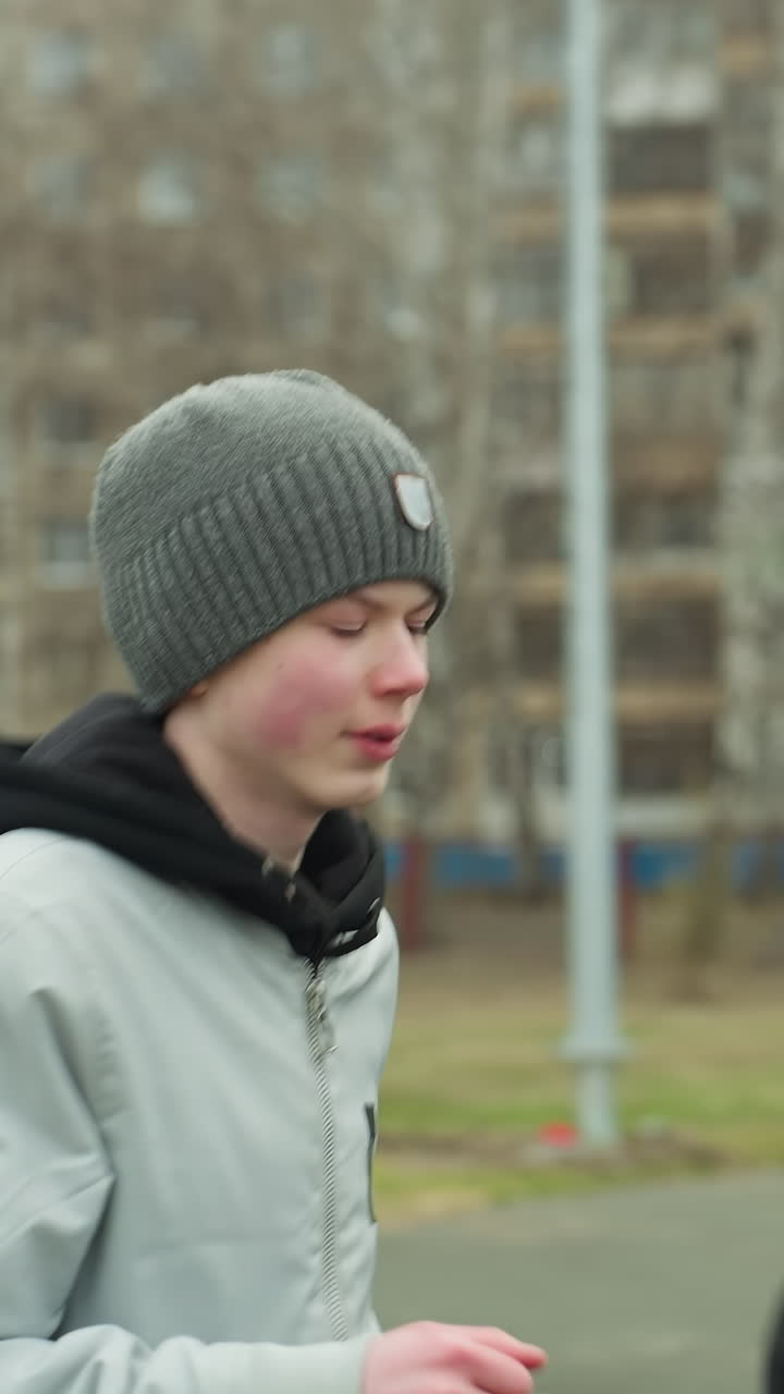 Close-up of two boys jogging gradually, with one wearing glasses and a gray beanie, as they jog, a man in a brown beanie speeds past them on the track, with a blurred background