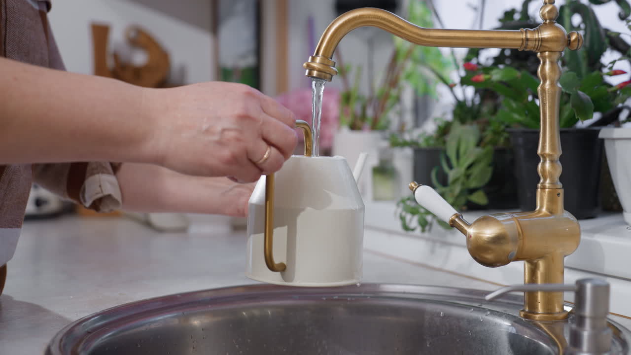 Botanical nurturer fetches water with white kettle from modern sink amid lush indoor plants and brass faucet detail in serene home gardening ritual on kitchen counter with flowing stream of water
