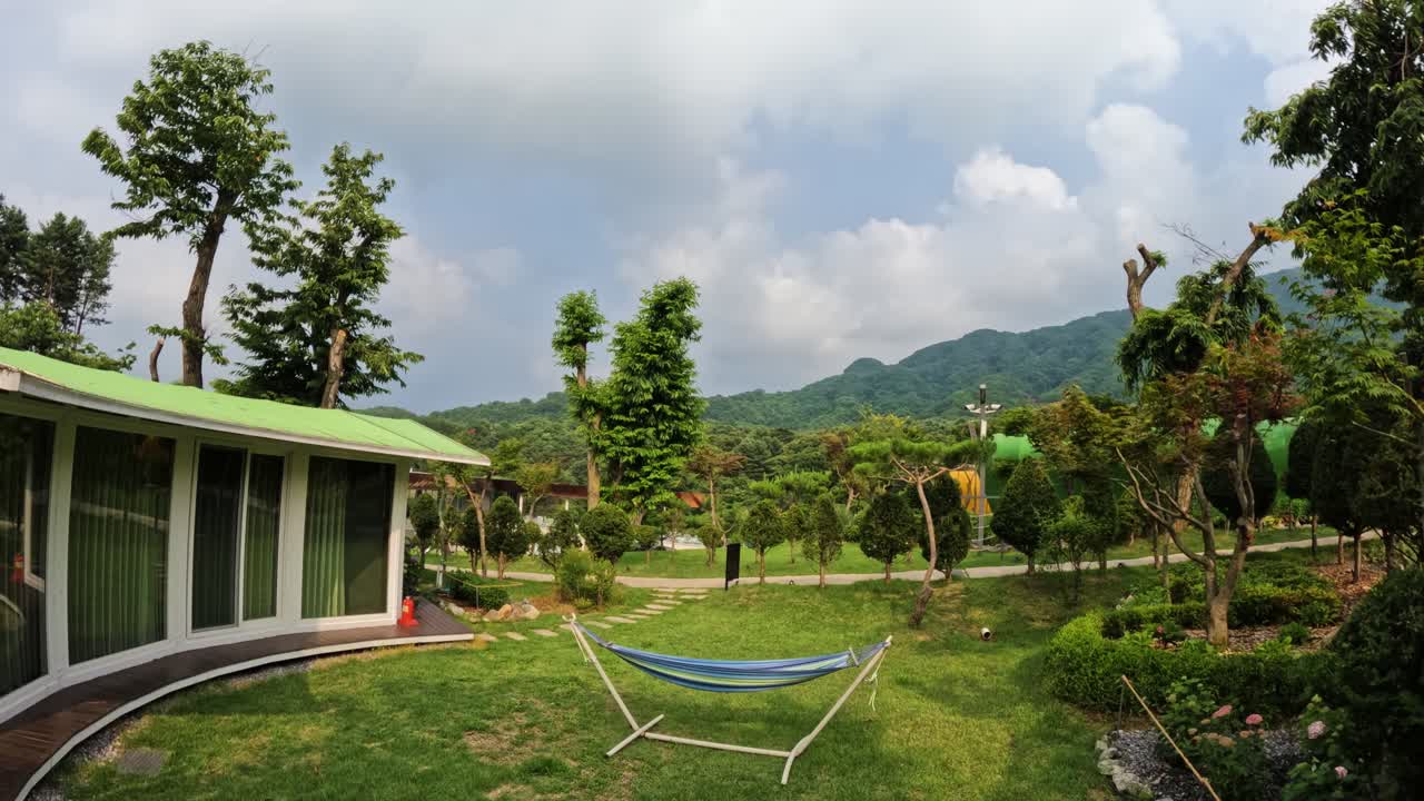 A scenic time-lapse captures fast-moving white clouds passing over a modern cabin, a relaxing hammock, and a lush green mountain landscape at a resort in Gapyeong, South Korea