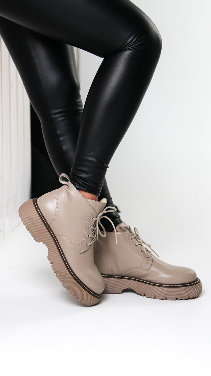 Woman in black leather pants and beige boots standing at the wall. Beige bag beside at the wall. White backdrop. Vertical video