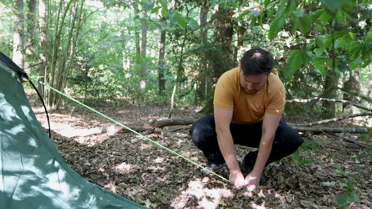 un hombre monta una tienda de campaña en un campamento en el bosque, pero no logra clavar el gancho en el suelo y deja caer la cuerda.