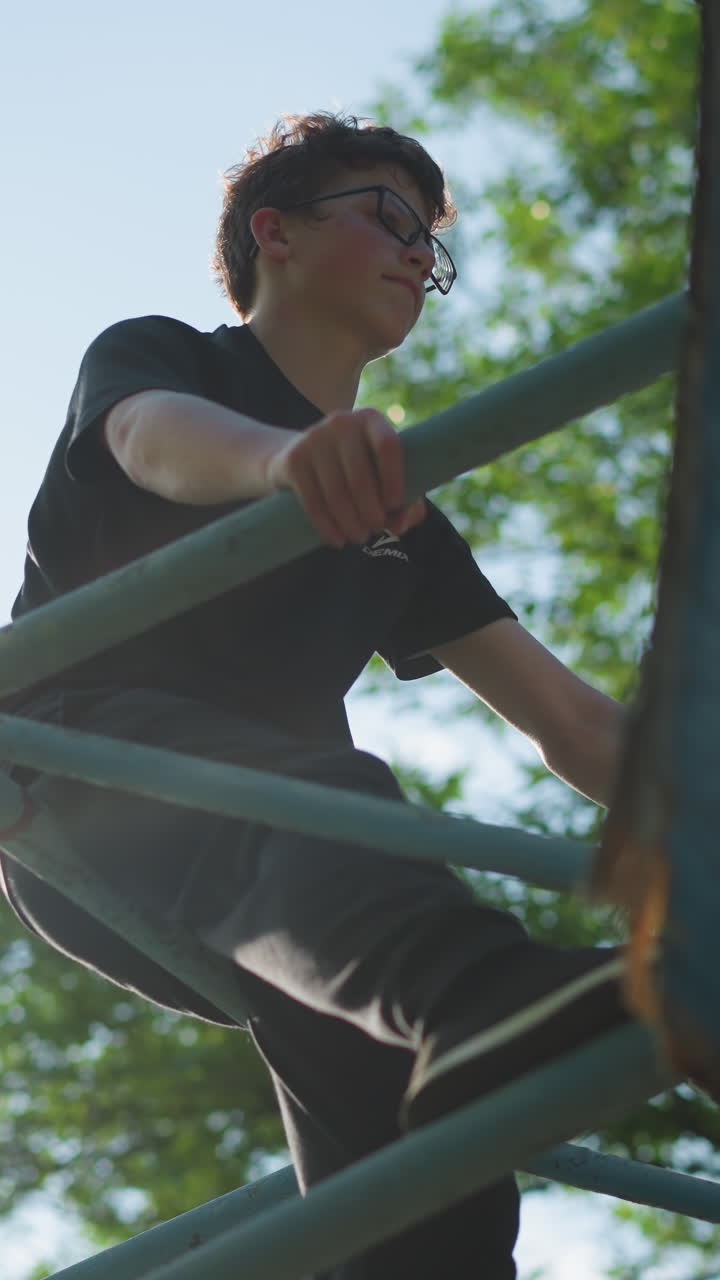 어린 소년은 야외의 파란 철 구조물에 앉아, 생각에 빠져, 두 손으로 프레임을 잡고, 빛은 그의 뒤에 있는 울창한 초록색 나무를 통해 필터링합니다.