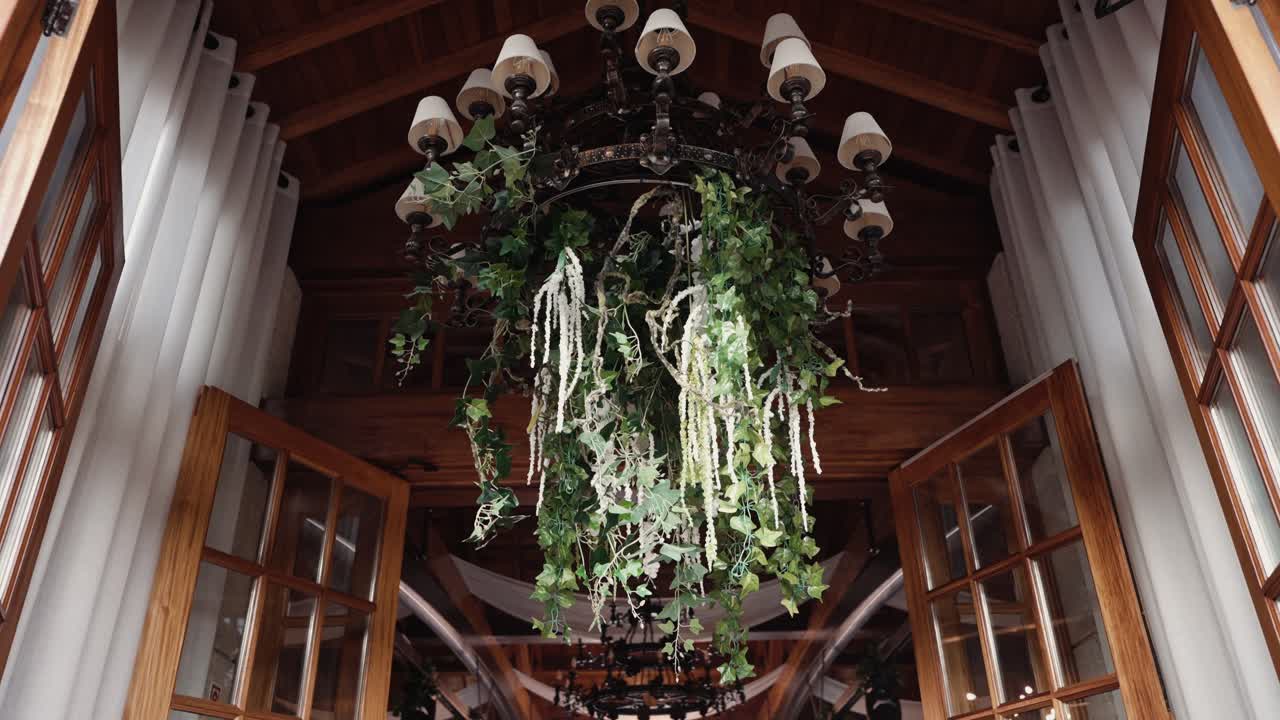 Lampadario decorativo con piante pendenti all'interno dell'ingresso di un edificio in legno con porte aperte.