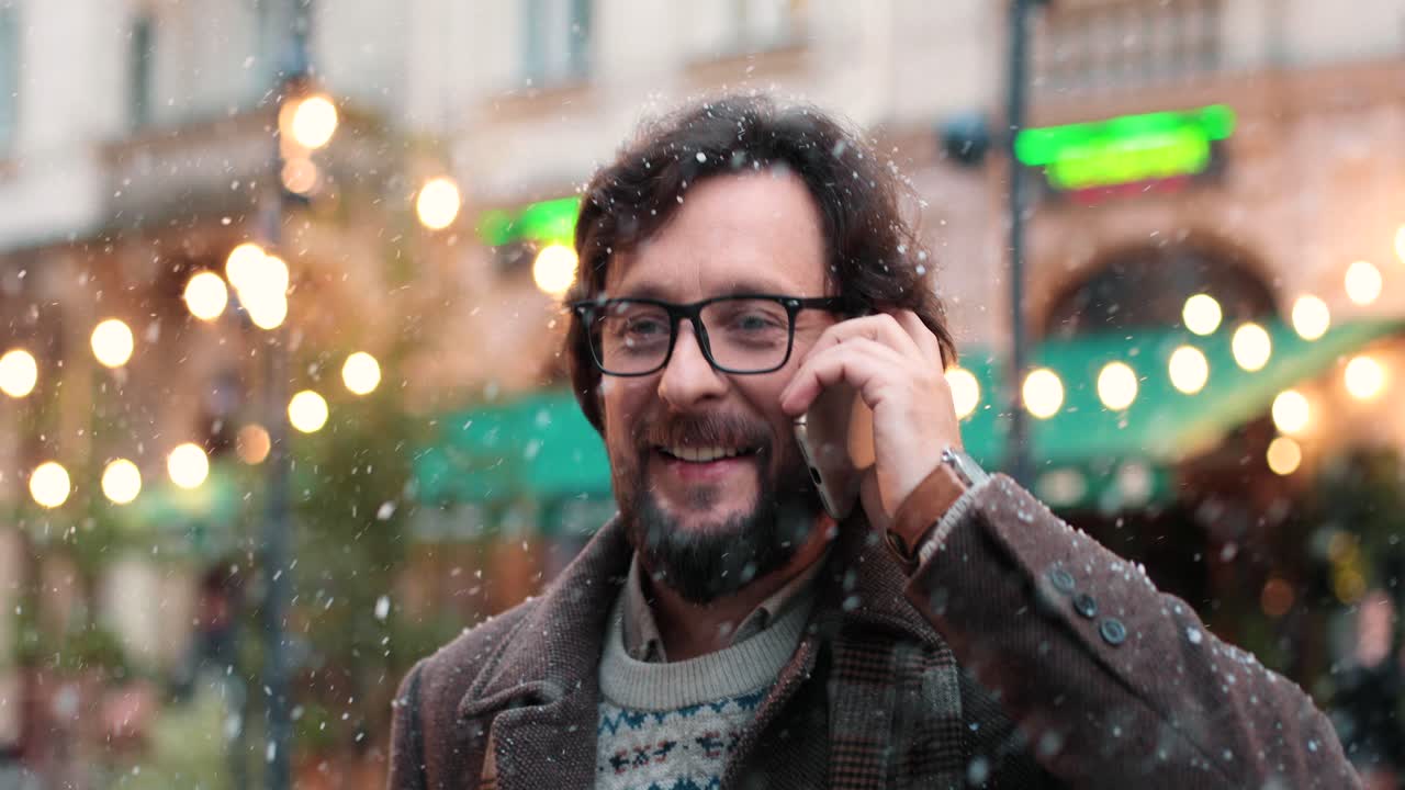 Close-up view of caucasian man wearing eyeglasses talking on smartphone on the street while it‚Äôs snowing in Christmas