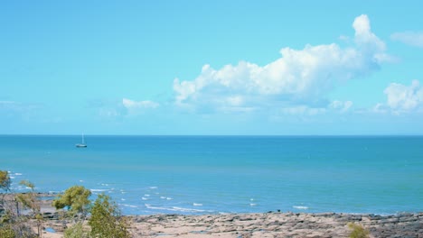 A medium sized sailing boat sits, floating upon tropical blue and green waters just off the coast of a rocky beach, looking out to the vast ocean that sits under the fluffy clouds floating above