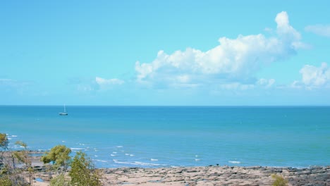 a medium sized sailing boat sits, floating upon tropical blue and green waters just off the coast of a rocky beach, looking out to the vast ocean that sits under the fluffy clouds floating above