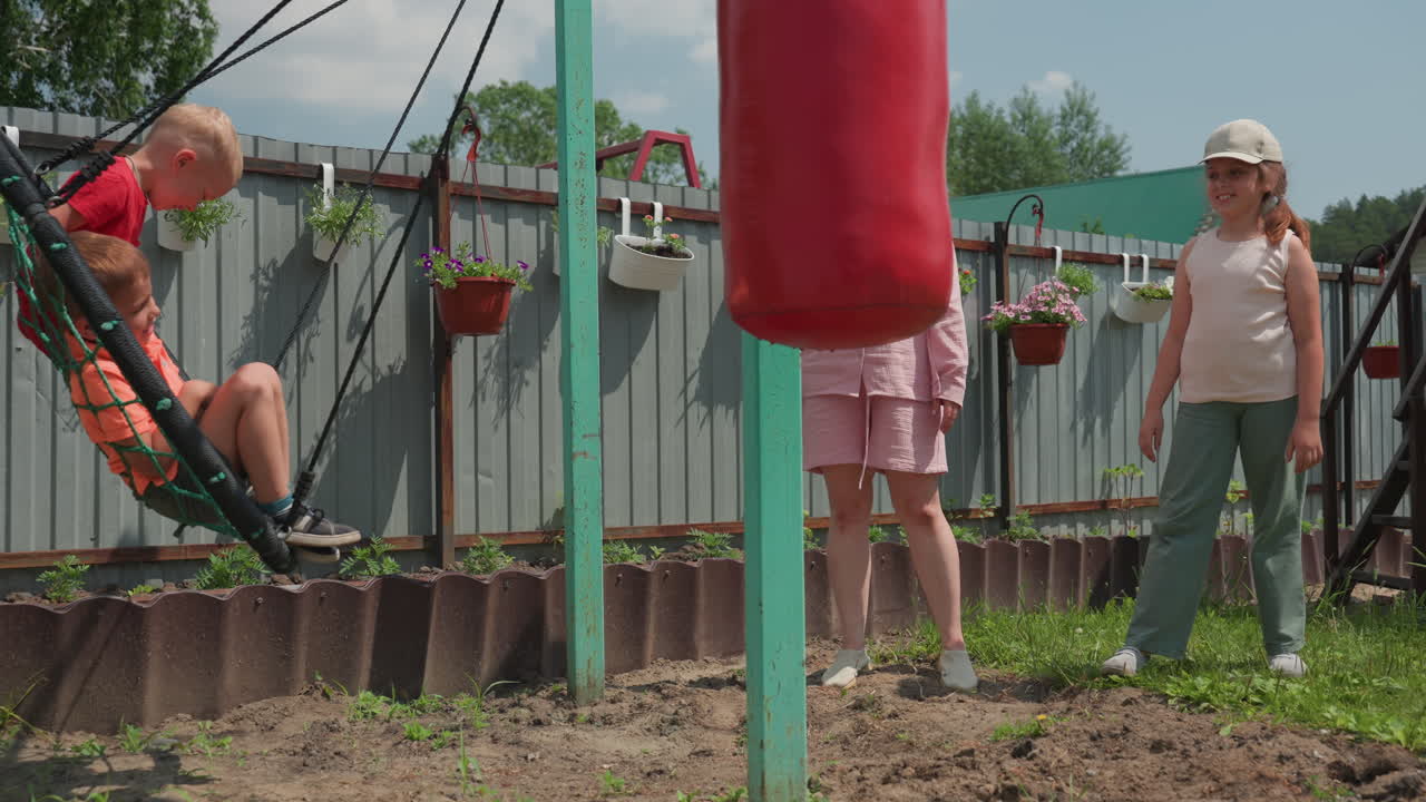 Caucasian Women Neighbors Jointly Pushing Children On Nest Swing In Backyard, Cooperative Caregiving Under Sunny Sky, Cheerful Interaction Between Adults And Kids Near Fence And Garden, Communal