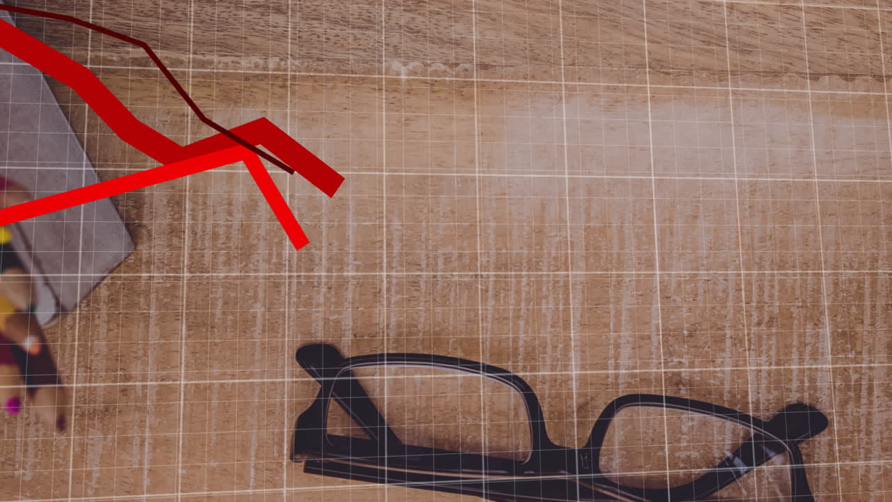 Black eyeglasses sitting on wooden desk, showing pencils, notebook with red trendline grid overlay