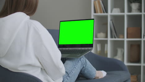 woman at home sitting on a couch works on a laptop computer with green mock-up screen. coronavirus covid-19 quarantine remote education or working concept. girl using computer browsing through