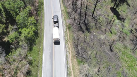aerial-of-a-truck-pulling-a-large-covered-trailer-down-a-mountain-road-with-burnt-dead-trees-surrounding-environment