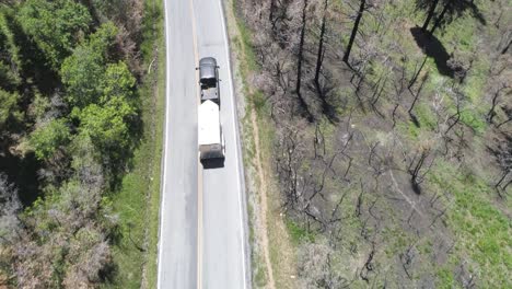 aerial of a truck pulling a large covered trailer down a mountain road with burnt-dead trees surrounding environment