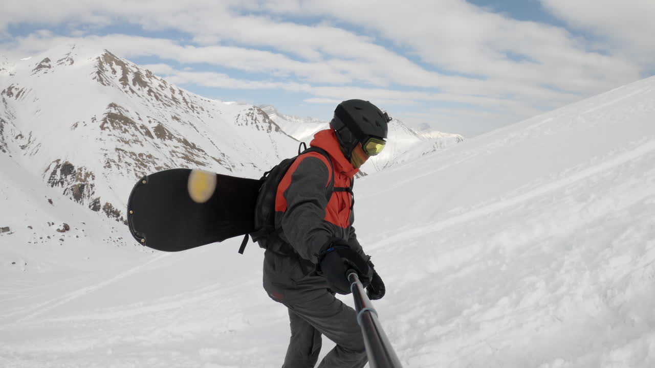 Young male freeride snowboarder walking up snow covered hill, Caucasus mountains, selfie