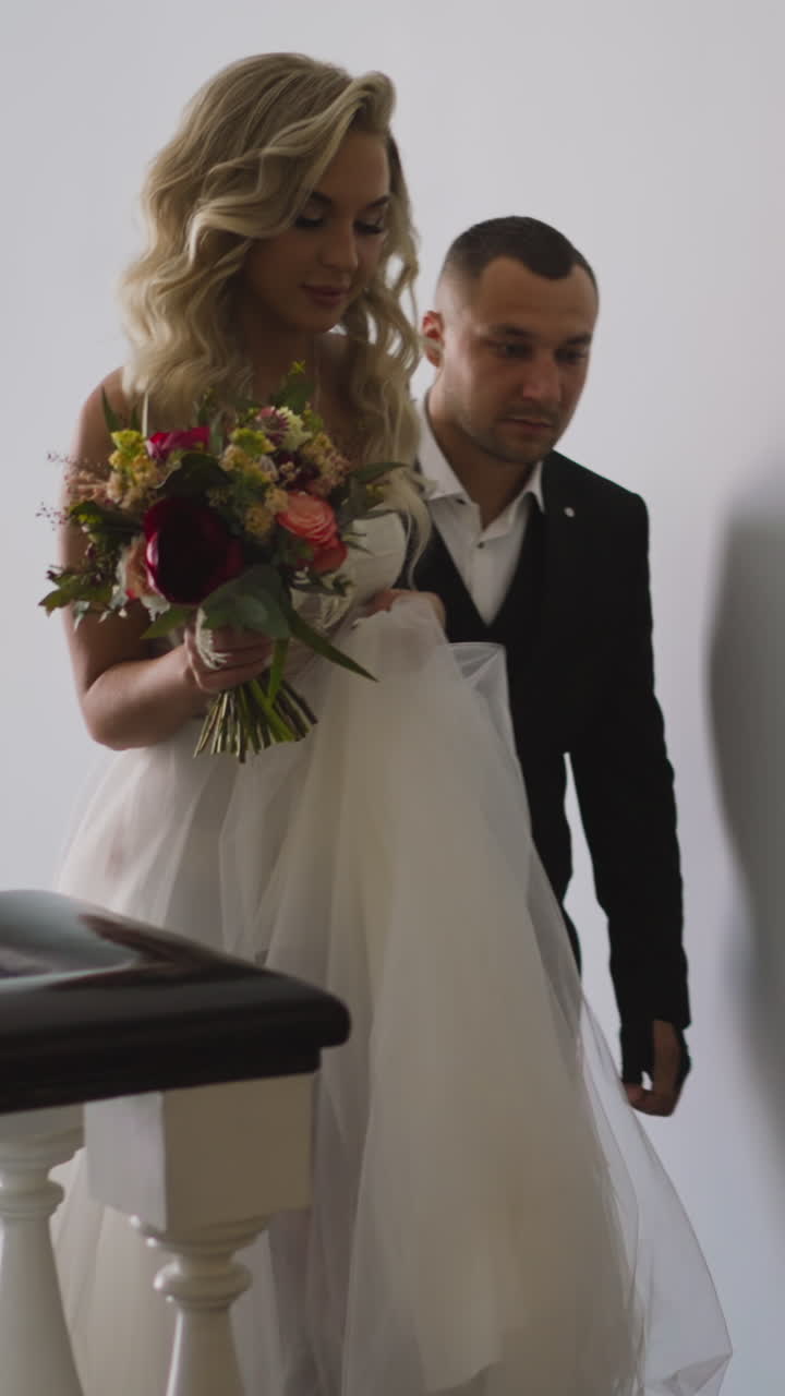 Pretty woman in wedding dress with hairstyle holding festive bouquet and groom in classic suit go up stairs in light hotel building hallway slow motion