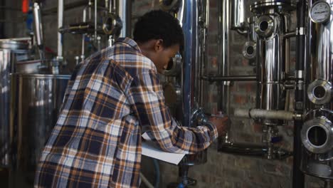 african american woman working at gin distillery checking equipment and writing on clipboard