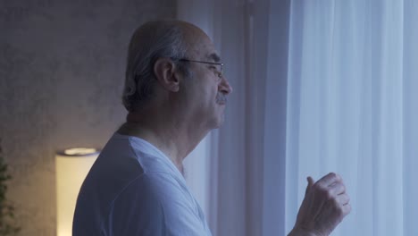 Unhappy-old-man-looking-out-the-window-in-the-living-room-at-home-takes-off-his-glasses.