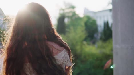 beautiful natural young girl with long red curly-wavy hair which is blown by wind, turning around and smiling, breathing the nature aromas, stroking her hair and looking somewhere