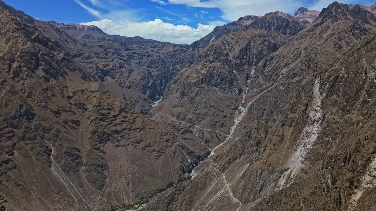 Um drone voa para dentro do cânion Colca, em suas profundezas duras e acidentadas. Paredes altíssimas se erguem em ambos os lados, criando uma paisagem dramática e vertical.