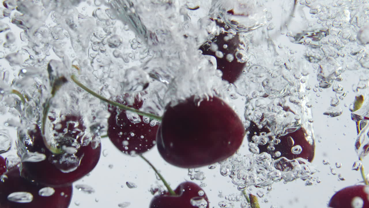 Primo piano di ciliegie mature che cadono nell'acqua creando schizzi e bollicine su sfondo bianco.