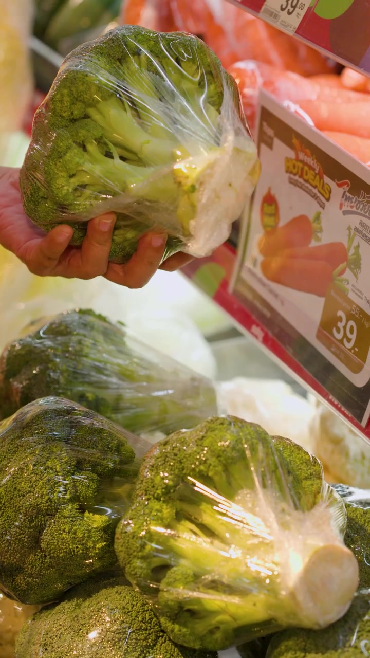 Primer plano de una mano recogiendo brócoli envuelto en plástico de un estante de supermercado en Bangkok.