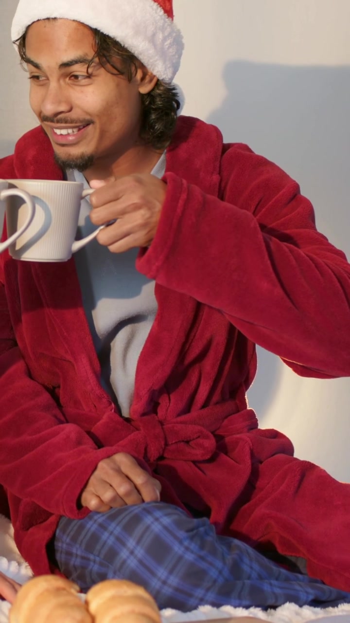 Vertical video: Seeing second white mug from left, man toasting with white mug on bed, in red robe