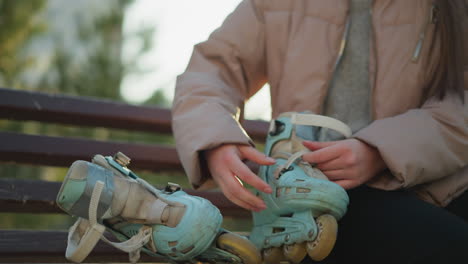 a person wearing a peach jacket is seated on a park bench, preparing to put on a pair of blue rollerblades. the close-up shot shows the individual s hands adjusting the rollerblades