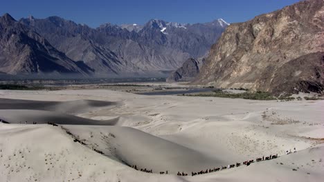 a dramatic desert scene featuring a line of camels and travelers traversing golden sand dunes, set against a backdrop of rugged, snow-capped mountains