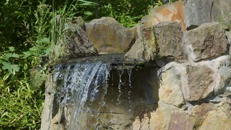 a steady stream of water cascades from a rustic stone fountain surrounded by greenery in a sunlit berlin park. static camera, natural daylight