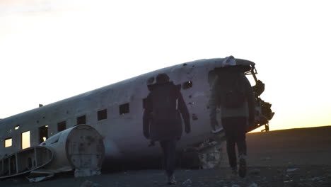 travelers walking towards crashed and abandoned airplane on black sand beach in sunset