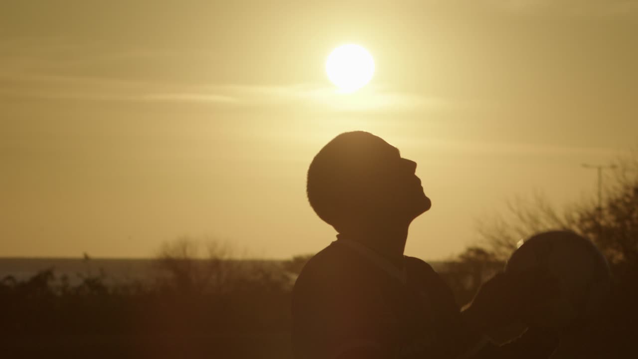 A person balances a football soccer ball on their forehead against a sunset sky in slow motion