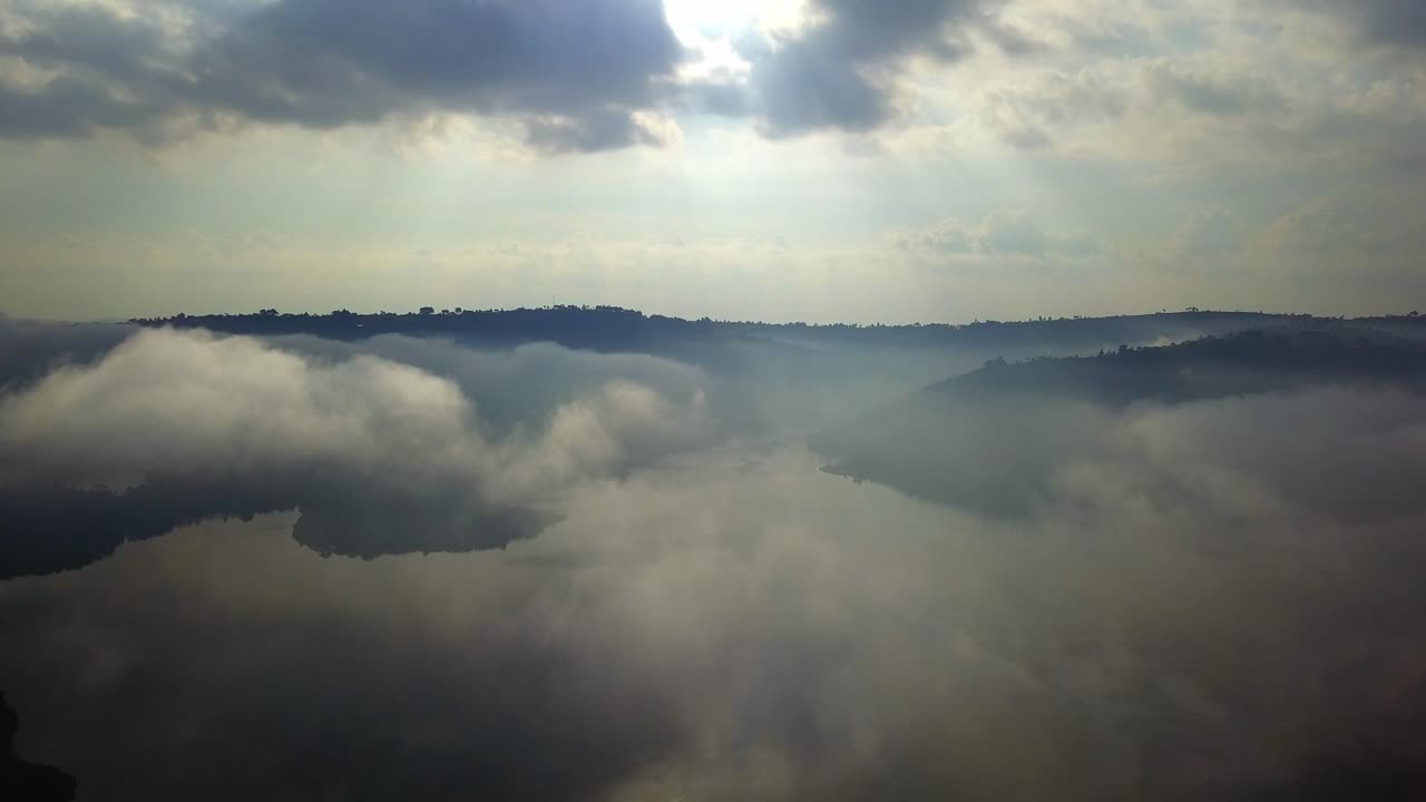 Drone view of mist and clouds rolling over the calm waters of Lake Bunyonyi in southwestern Uganda, Africa, with soft sunlight filtering through and green hills reflecting on the peaceful lake surface