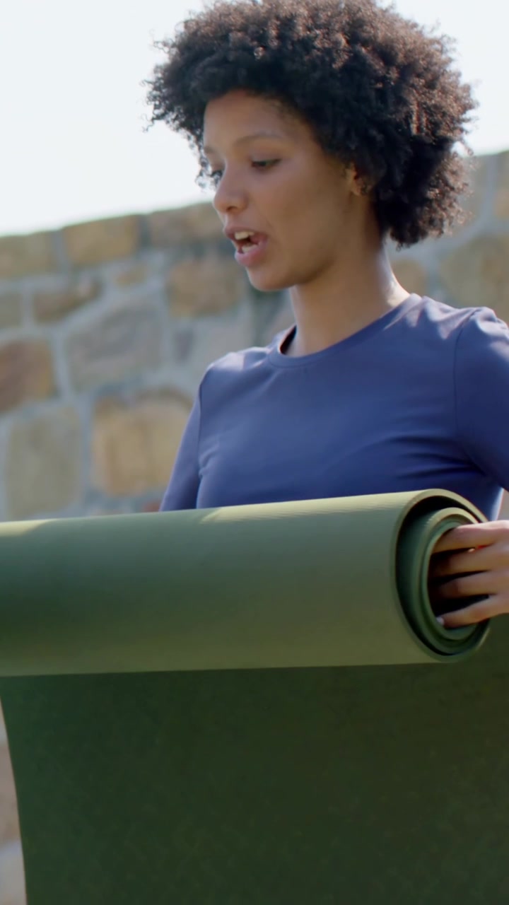 Video verticale: Donna in blu scuro che prepara un tappetino verde e regola la presa nel patio per lo yoga, con smartwatch.
