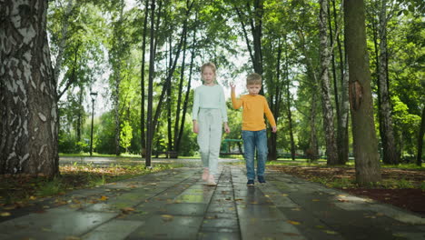 Young boy and girl dressed in stylish outfits walk hand in hand through sunny park pathway surrounded by green trees, sharing joyful moment and casual gestures on peaceful autumn day outdoors