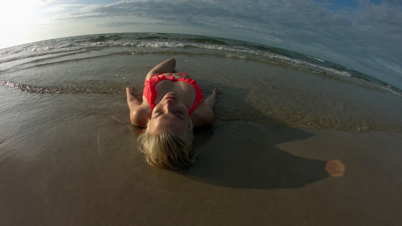 Shallow ocean waves going over a woman wearing an orange bikini, using a fish eye lens