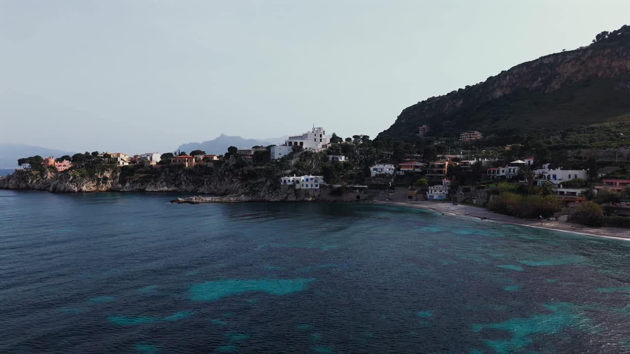 Drone flies steadily toward coastal cliffs and colorful buildings in Porticello, Sicily, capturing deep blue water, seaside houses, and mountain ridges in northern Sicily. Palermo