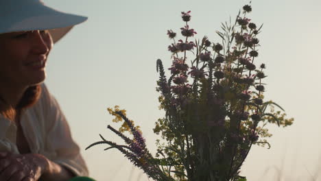 woman in white sunhat smiling while admiring wildflower bouquet held against sunlight with soft golden rays glowing through petals and stems