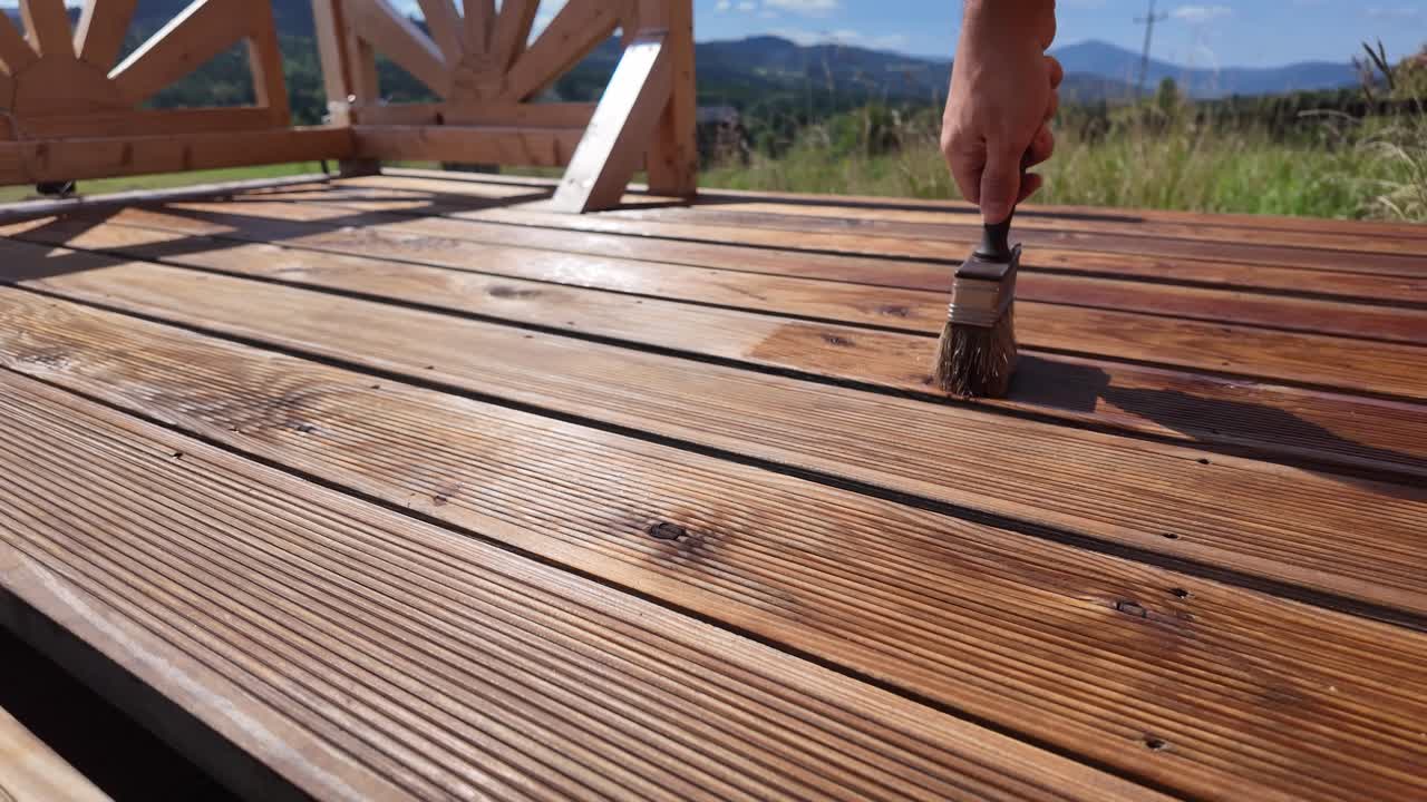 Hand oils varnishing impregnating wooden terrace boards with brush. Decorative railing, serene sky, clouds, and mountain horizon evoke home improvement and outdoor maintenance themes