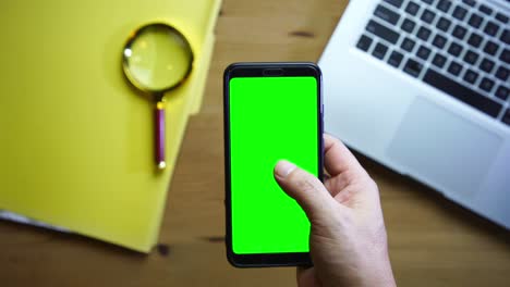 man holding green screen smart phone  against laptop and magnifying glass