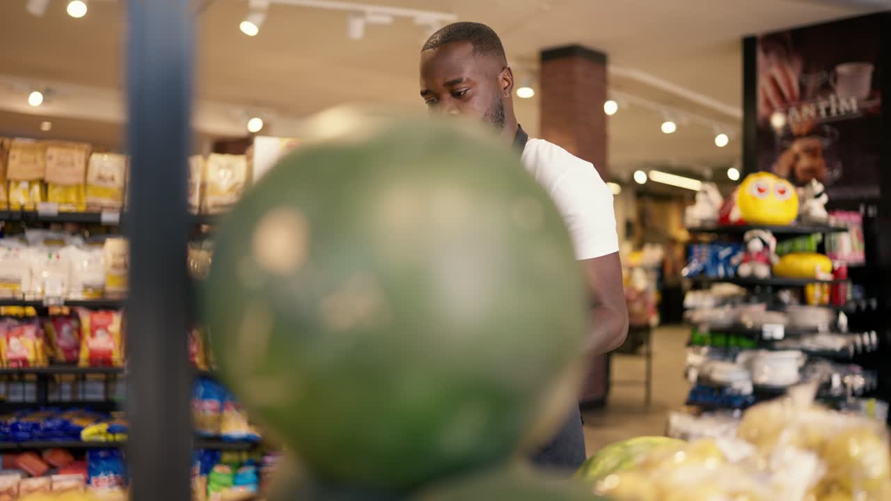 En mørkhudet mann i en hvit T-skjorte og svart forkle bærer en vannmelon og legger den på supermarkedets disk. Supermarked om sommeren