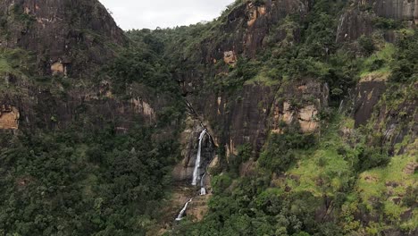 aerial far shot of deep waterfall flowing smoothly between green huge mountains, sri lanka