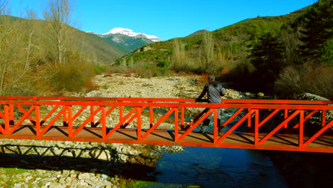 Vista-Aérea-De-Un-Hombre-Parado-En-Un-Puente-Rojo-De-Montaña-Contemplando-El-Paisaje-De-Los-Pirineos-Españoles