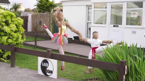 Dressed-Scarecrows-In-An-English-Village-In-The-United-Kingdom-With-Lockdown-Sign-In-Yin-Yang-Icon-On-The-Fence-During-Covid-19-Pandemic---full-shot