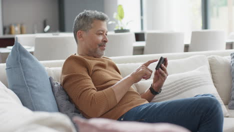 Happy-senior-biracial-man-using-smartphone-on-couch-in-sunny-living-room,-slow-motion