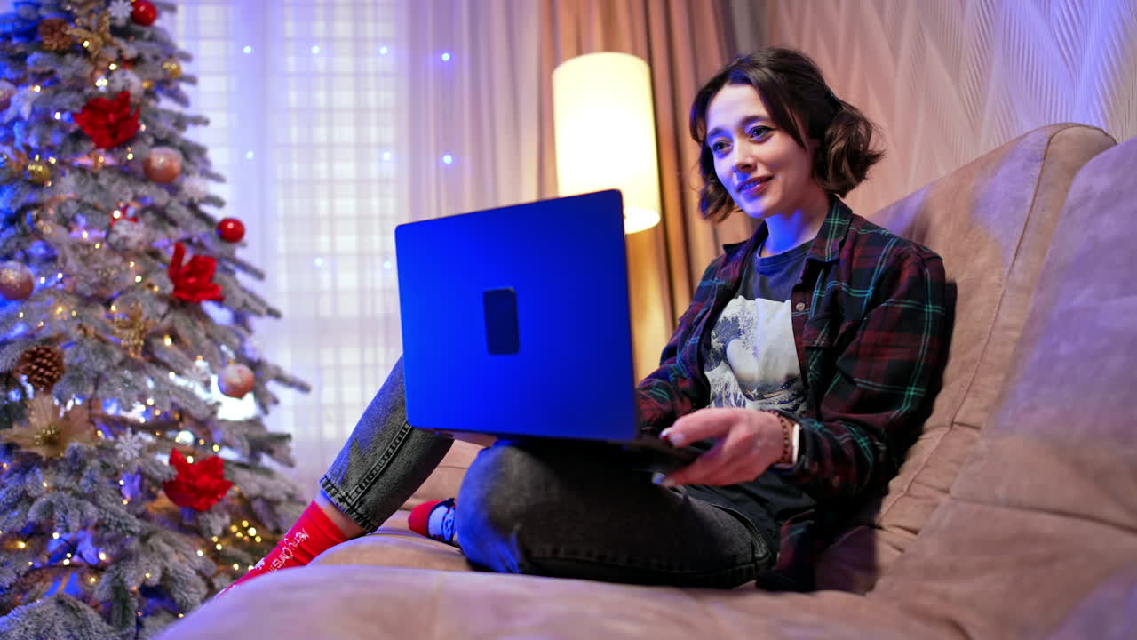 Working from home during the holidays. A person engages with their laptop while sitting on a couch near a decorated Christmas tree in cozy evening lighting