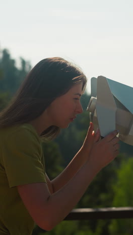 curious woman relishes view with interest and looks through binoculars standing on observation deck. tourist area surrounded by thick coniferous woodland